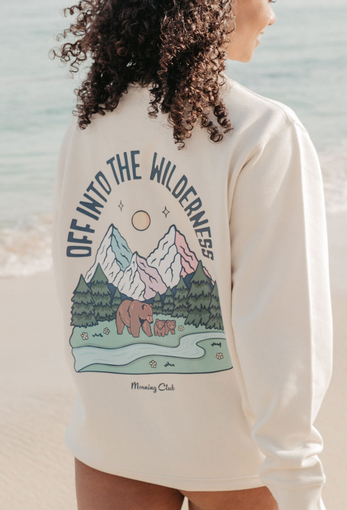 Lady wearing a printed sweatshirt, that says into the wilderness, and its set on the beach.