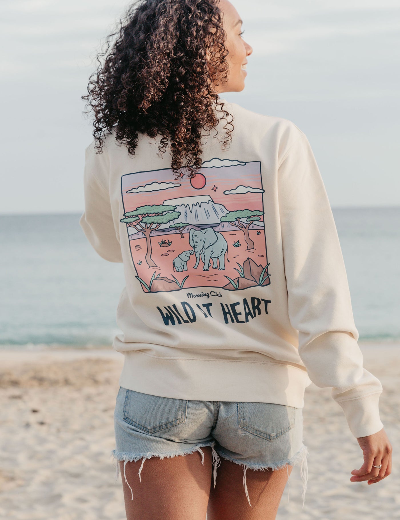 Lady wearing a printed sweatshirt, that says wild at heart, and its set on the beach.