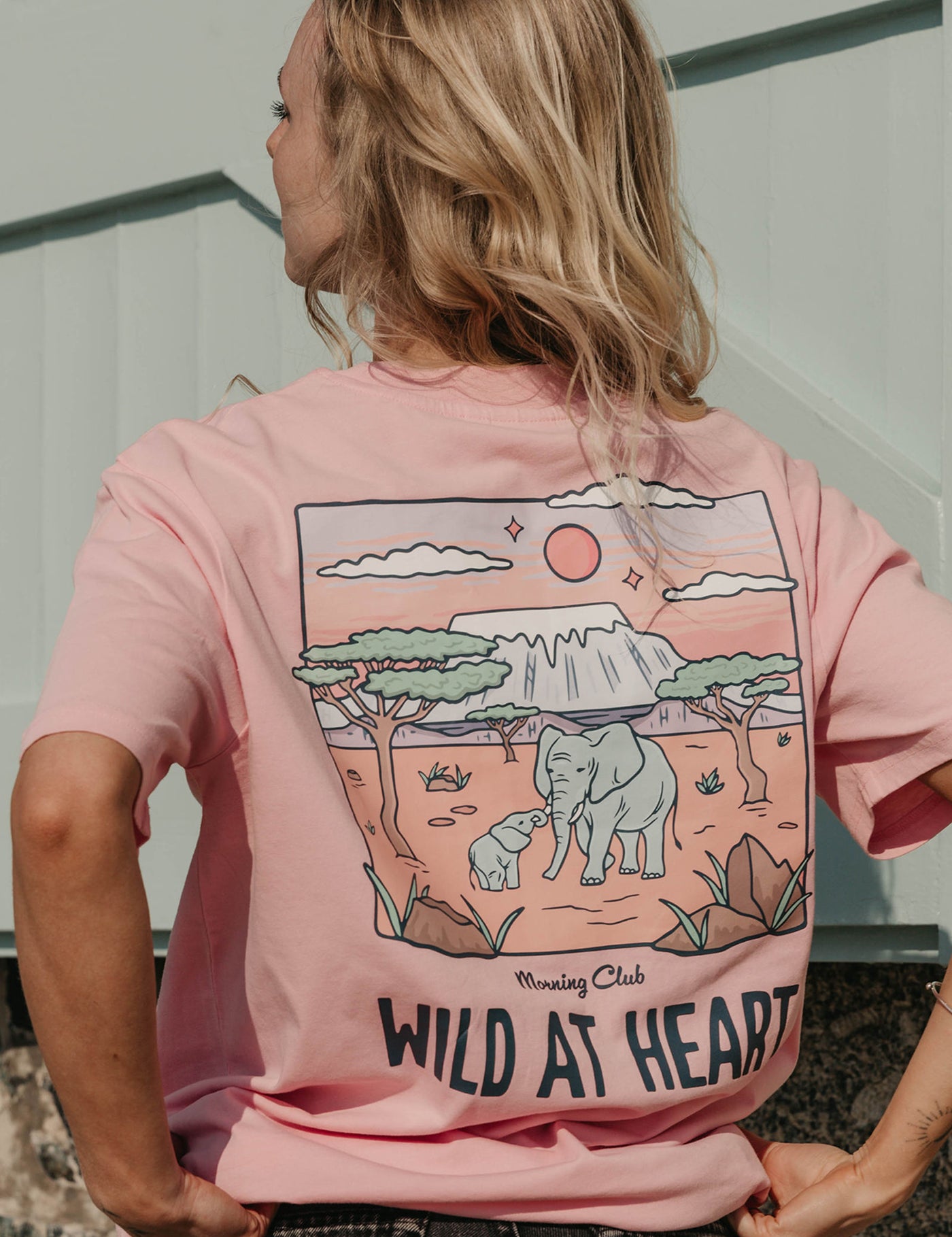 A lady wearing a pink printed graphic t-shirt, that says wild at heart, set by the beach.