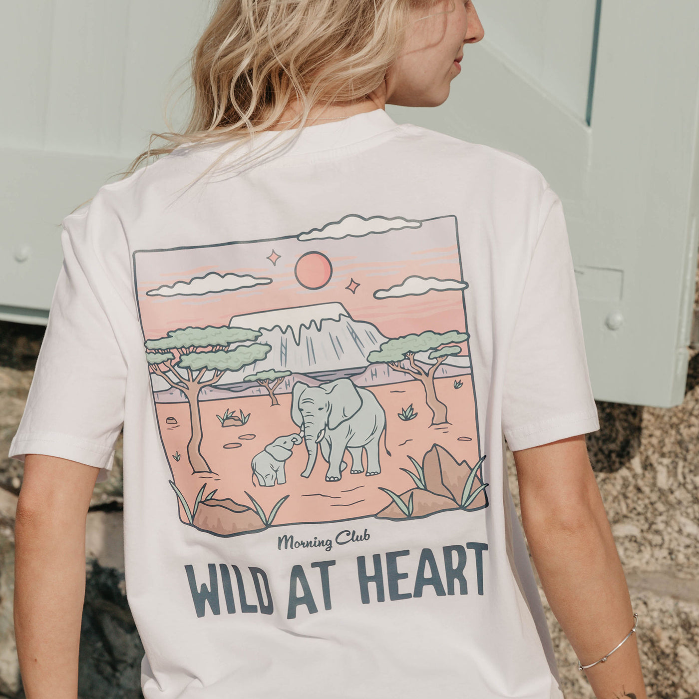 A lady wearing a white printed graphic t-shirt, that says wild at heart, set by the beach.