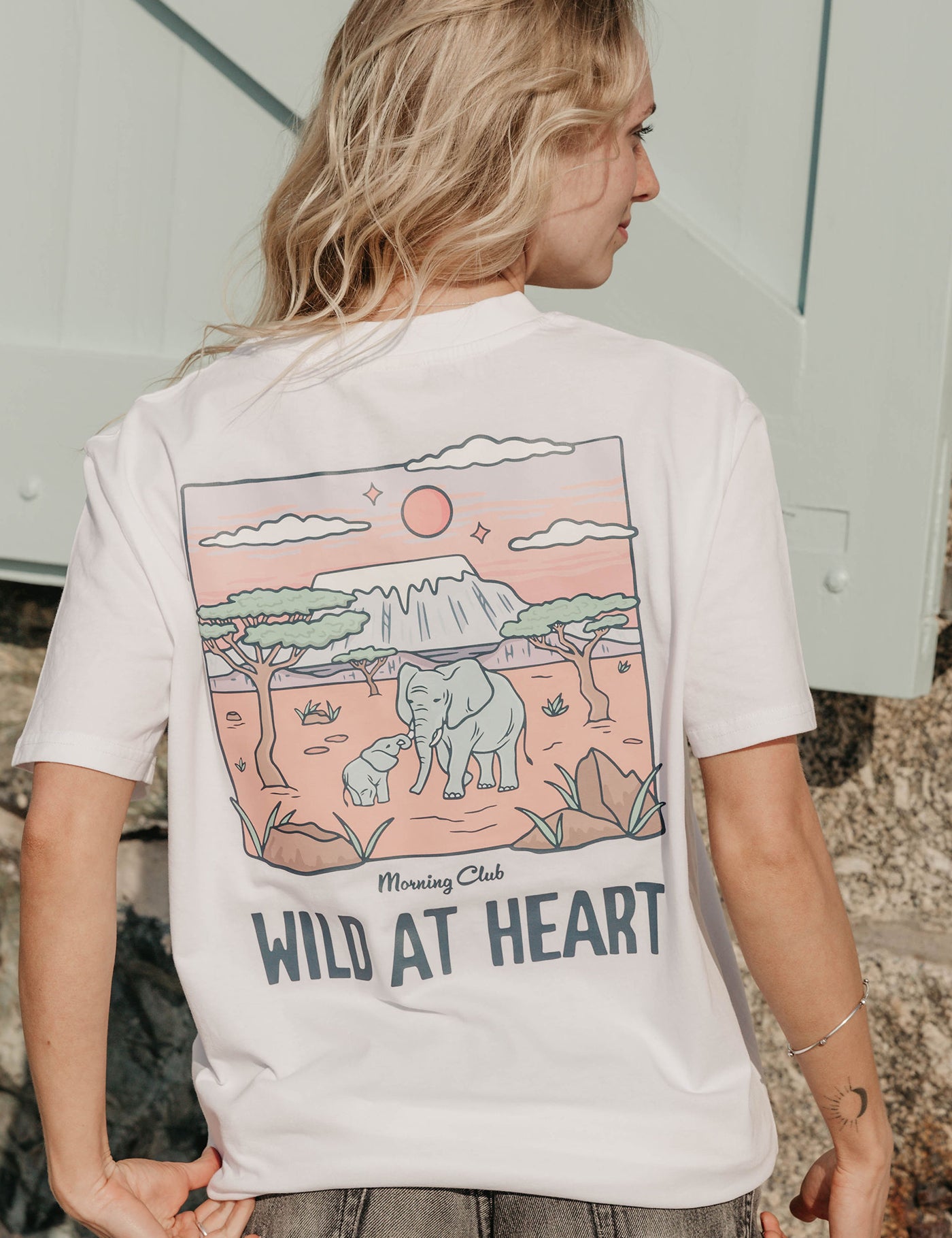 A lady wearing a white printed graphic t-shirt, that says wild at heart, set by the beach.